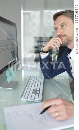 Businessman in a blue jacket working on computer, typing and writing notes in modern office, managing tasks, multitasking. Business people concept Businessman in a blue jacket working on computer, typing and writing notes in modern office, managing tasks, multitasking. Business people concept 137716542