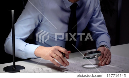 Businessman diligently auditing financial documents with a magnifying glass under focused lamp light. Audit and taxes in business Businessman diligently auditing financial documents with a magnifying glass under focused lamp light. Audit and taxes in business 137716545