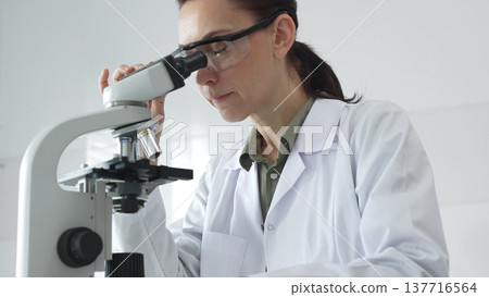 Female scientist in a lab coat and protective glasses examining samples through a microscope in a research laboratory. Medicine, healthcare and science concept 137716564
