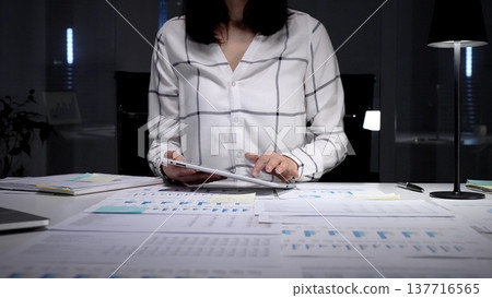 Businesswoman working late night in a modern office, analyzing financial data on a digital tablet and reviewing paperwork, demonstrating dedication and overtime productivity Businesswoman working late night in a modern office, analyzing financial data on a digital tablet and reviewing paperwork, demonstrating dedication and overtime productivity 137716565