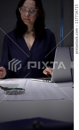 Woman working late night in office, multitasking with a tablet and writing on documents, managing heavy paperwork load. Audit and taxes in business Woman working late night in office, multitasking with a tablet and writing on documents, managing heavy paperwork load. Audit and taxes in business 137716715