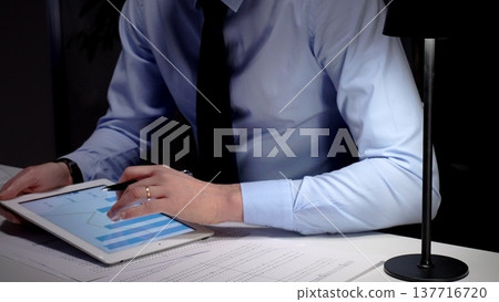 Businessman working late analyzing financial data at office, holding tablet with charts, writing notes on documents. Audit and taxes in business Businessman working late analyzing financial data at office, holding tablet with charts, writing notes on documents. Audit and taxes in business 137716720