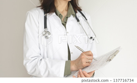 Female doctor, wearing a stethoscope and a medical coat over a green blouse, is standing and writing on a white clipboard, representing medical expertise and patient care. Medicine concept. Female doctor, wearing a stethoscope and a medical coat over a green blouse, is standing and writing on a white clipboard, representing medical expertise and patient care. Medicine concept. 137716734