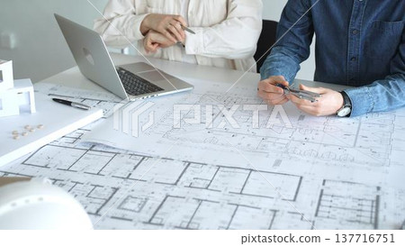 Engineering professionals collaborating on an architectural blueprint, using a compass to design a modern building, planning construction with a house model and laptop 137716751