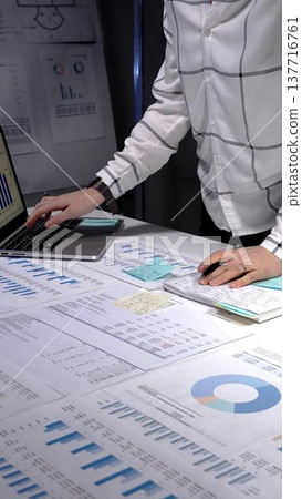 Business professional analyzing financial data and statistics. Working late night in a modern office. Using a laptop and reviewing documents with charts and graphs 137716761