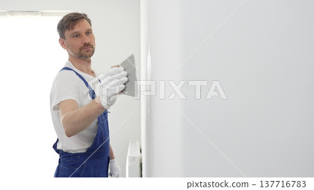 Middle aged man construction worker wearing gloves and blue overalls is using a putty knife to apply plaster on a white wall, performing home renovation and improvement Middle aged man construction worker wearing gloves and blue overalls is using a putty knife to apply plaster on a white wall, performing home renovation and improvement 137716783