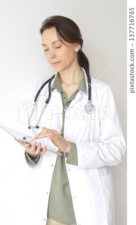 Portrait of a doctor woman wearing white medical coat over green blouse, stethoscope, is reviewing patient records on digital tablet, standing in medical workspace with natural lighting. Medicine 137716785