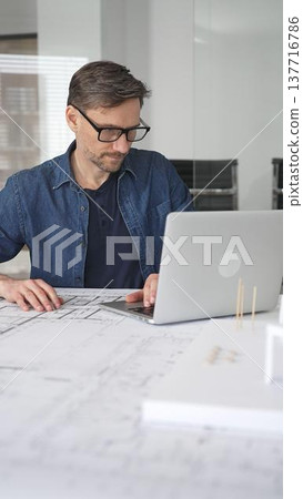 Male architect in glasses at a contemporary office working on a white architectural scale model of a modern house design, developing creative housing plans on a desk with blueprints and a laptop 137716786