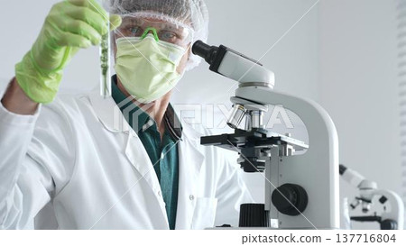 Adult male scientist wearing green protective gear looking at plant samples in test tubes and using a microscope in modern laboratory for botanical research. Medicine, healthcare and science concept 137716804