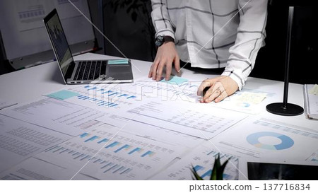 Professional woman reviewing financial reports, analyzing market data, and developing business strategy using a pen, laptop, and extensive paperwork on an office desk at night Professional woman reviewing financial reports, analyzing market data, and developing business strategy using a pen, laptop, and extensive paperwork on an office desk at night 137716834