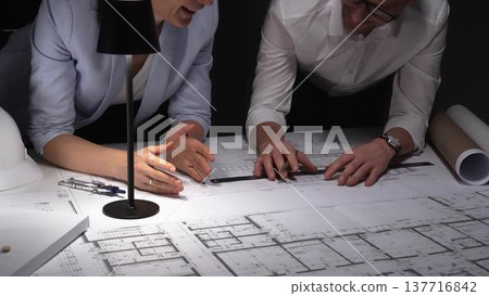 Architects collaborating on blueprints, diligently planning building design and construction project details late at night. Architects collaborating on blueprints, diligently planning building design and construction project details late at night. 137716842