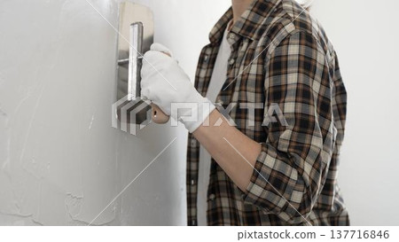 Unknown female construction worker wearing protective gloves and checkered shirt is smoothing wet plaster across white wall, spreading material with professional trowel during interior renovation Unknown female construction worker wearing protective gloves and checkered shirt is smoothing wet plaster across white wall, spreading material with professional trowel during interior renovation 137716846