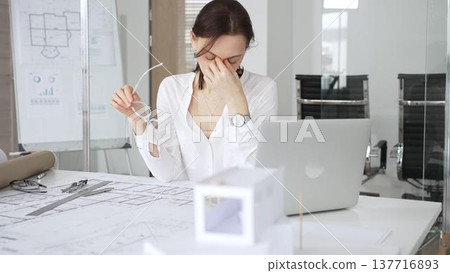 Professional architect woman working on architectural plans. Finding inspiration for a new building project. Smiling with satisfaction and confidence in her modern office Professional architect woman working on architectural plans. Finding inspiration for a new building project. Smiling with satisfaction and confidence in her modern office 137716893