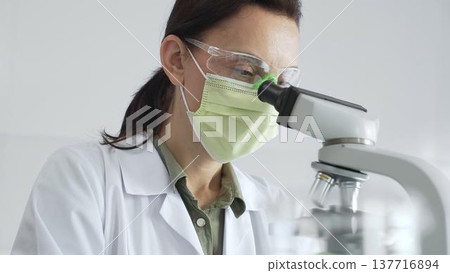 Female professional scientist examining specimen, placing slide under microscope while wearing green protective gear in laboratory workspace. Medicine, health care and science concept Female professional scientist examining specimen, placing slide under microscope while wearing green protective gear in laboratory workspace. Medicine, health care and science concept 137716894