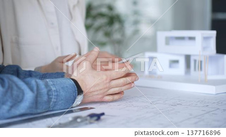 Architects working on blueprint and architectural model, hands detailing building plans with a compass, discussing design and construction project development Architects working on blueprint and architectural model, hands detailing building plans with a compass, discussing design and construction project development 137716896