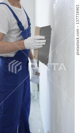 Man construction worker wearing protective gloves and blue construction coveralls, spreading plaster smoothly across wall using professional drywall taping knife Man construction worker wearing protective gloves and blue construction coveralls, spreading plaster smoothly across wall using professional drywall taping knife 137716901