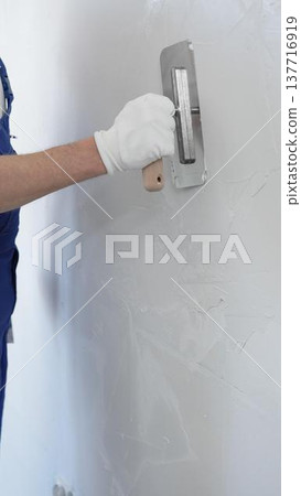 Professional construction worker wearing blue work overall is using a finishing trowel to carefully apply plaster on a wall, demonstrating expertise in home renovation Professional construction worker wearing blue work overall is using a finishing trowel to carefully apply plaster on a wall, demonstrating expertise in home renovation 137716919