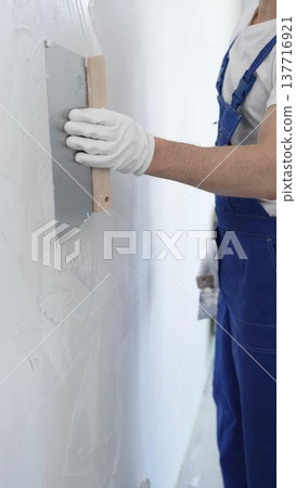 Male construction worker wearing protective gloves and blue construction coveralls, spreading plaster smoothly across wall using professional drywall taping knife during renovation, vertical view Male construction worker wearing protective gloves and blue construction coveralls, spreading plaster smoothly across wall using professional drywall taping knife during renovation, vertical view 137716921