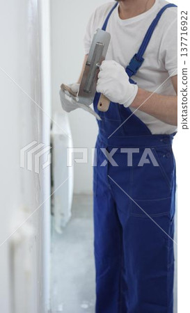 Professional construction worker wearing blue work overall is using a finishing trowel to carefully apply plaster on a wall, demonstrating expertise in home renovation Professional construction worker wearing blue work overall is using a finishing trowel to carefully apply plaster on a wall, demonstrating expertise in home renovation 137716922