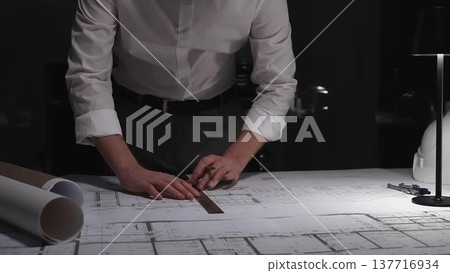 Architect working on night shift, drawing blueprints with precision ruler for new construction project design. 137716934