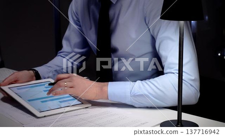 Businessman working late analyzing financial data at office, holding tablet with charts, writing notes on documents. Audit and taxes in business Businessman working late analyzing financial data at office, holding tablet with charts, writing notes on documents. Audit and taxes in business 137716942