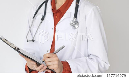 Unknown woman doctor wearing stethoscope and white coat over brick red blouse is standing and writing on a clipboard, representing medical expertise and patient care. Medicine and health care concept Unknown woman doctor wearing stethoscope and white coat over brick red blouse is standing and writing on a clipboard, representing medical expertise and patient care. Medicine and health care concept 137716979