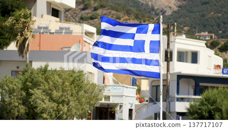 View of a Greek flag waving in front of buildings in a coastal town in Greece during a sunny day 137717107