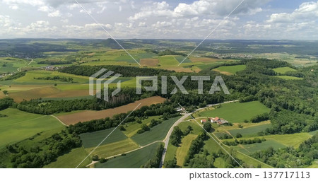 Green fields and hills stretch across the countryside with a winding road and small houses below in summer light Green fields and hills stretch across the countryside with a winding road and small houses below in summer light 137717113