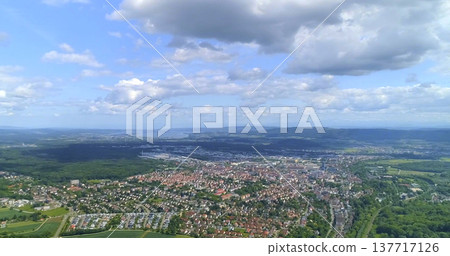 View of a city and land from above showing buildings, roads, and green areas during a sunny day in summer with clouds in the sky 137717126