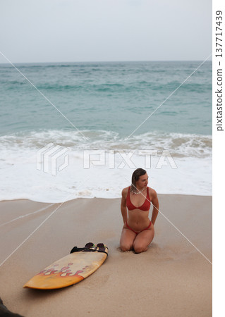 Woman Awaits Surf. Kneeling Lady Observes Ocean Waves. Contemplative Woman On Beach With Surfboard Woman Awaits Surf. Kneeling Lady Observes Ocean Waves. Contemplative Woman On Beach With Surfboard 137717439