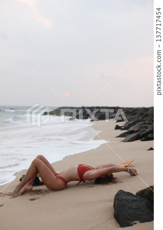 Tranquil Female Individual Reclining On Moist Sandy Beach Overlooking Gentle Ocean Waves At Dusk 137717454