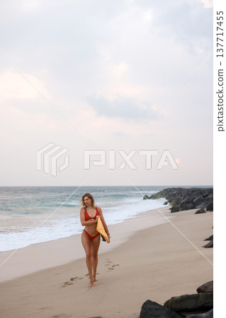 Serene Scene Of Female Surfer Strolling Along Sandy Coast With Yellow Board Under Cloudy Sky 137717455