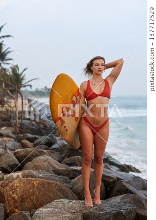 Graceful Woman With Surfboard At Rocky Coast. Selfassured Woman In Swimwear By Rugged Shoreline 137717529