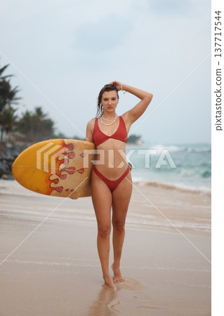 Elegant Female Model Posing In Red Bikini Along Tropical Shoreline With Layered Beads And Gentle Waves Elegant Female Model Posing In Red Bikini Along Tropical Shoreline With Layered Beads And Gentle Waves 137717544