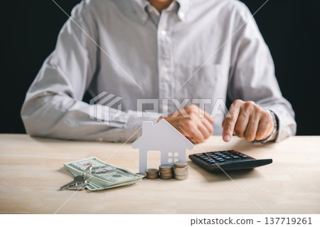 Person calculates home interest rate for mortgage loan, evaluating property investment money using calculator, stacks coins, and house model with keys on wooden table for real estate planning 137719261