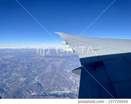 A view from an airplane window: the wing of an aircraft flying over a blue sky and snow-capped mountains. 137720056