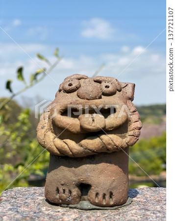 A humorous unglazed Shisa statue with its tongue sticking out against a blue sky, set against the backdrop of Okinawa. A humorous unglazed Shisa statue with its tongue sticking out against a blue sky, set against the backdrop of Okinawa. 137721107