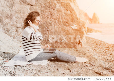 Freelancer working laptop woman sitting on pebble beach by the ocean during sunset with remote work concept Freelancer working laptop woman sitting on pebble beach by the ocean during sunset with remote work concept 137722333