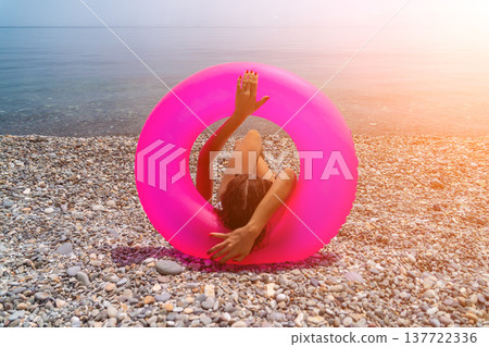 Beach Woman Inflatable sitting inside a large pink ring on a pebble shore enjoying a peaceful summer sunset Beach Woman Inflatable sitting inside a large pink ring on a pebble shore enjoying a peaceful summer sunset 137722336
