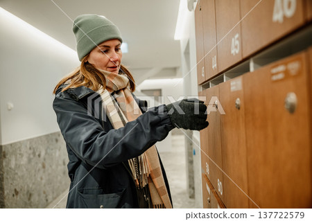Woman checks mail in a modern building hallway wearing winter clothes during day time Woman checks mail in a modern building hallway wearing winter clothes during day time 137722579