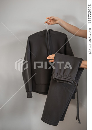 A person holds a black zip-up sweater and matching pants in a simple indoor setting during daytime A person holds a black zip-up sweater and matching pants in a simple indoor setting during daytime 137722608