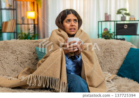 Indian woman drinking hot tea and coughing at home under blanket, showing cold and tired expression 137725394
