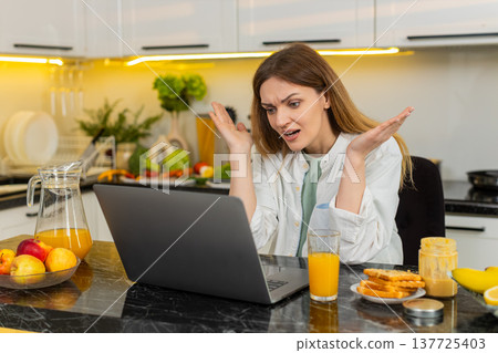 Middle-aged woman shocked by bad news on laptop in kitchen feeling stress after failure loss online Middle-aged woman shocked by bad news on laptop in kitchen feeling stress after failure loss online 137725403