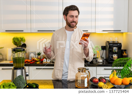 Middle-aged man drinks water checking smartphone in kitchen for weight loss diet hydration concept Middle-aged man drinks water checking smartphone in kitchen for weight loss diet hydration concept 137725404
