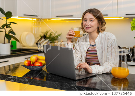 Young woman drinking orange juice while using laptop in kitchen for healthy eating and online chat 137725446