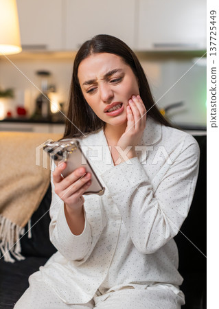 Young woman holds cheek tooth pain searches dentist on smartphone books visit dental care at night 137725449