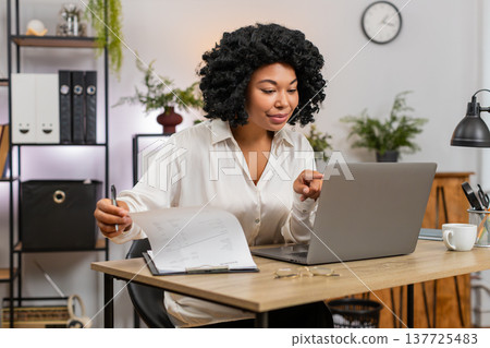African American businesswoman reviews paperwork nods satisfied seeing laptop data confirm records African American businesswoman reviews paperwork nods satisfied seeing laptop data confirm records 137725483