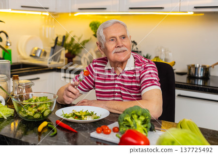 Senior old man happily eating fresh vegetable salad in home kitchen for healthy diet and weight loss 137725492