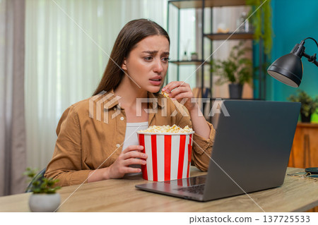Young woman watching dramatic movie on laptop eating popcorn nervously reacting to intense scene Young woman watching dramatic movie on laptop eating popcorn nervously reacting to intense scene 137725533