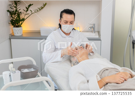 Beautician cosmetologist massaging peeling serum on middle aged young adult woman face in clinic 137725535